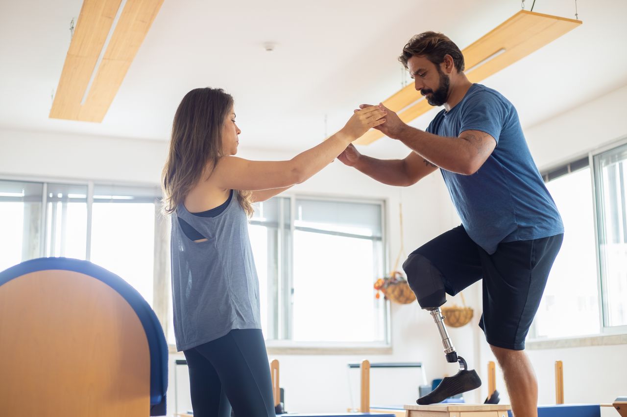A man with a prosthetic leg exercises with a therapist in a well-lit gym environment.